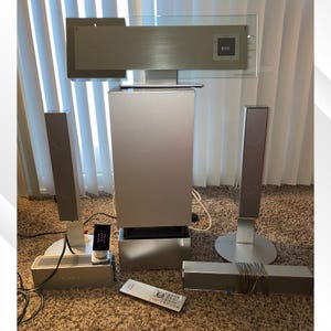 May include: A silver home theater system with a DVD player, subwoofer, two tall speakers, and a soundbar. The system includes a remote control and a small control panel. The speakers are on white stands, and the system is on a brown carpet.