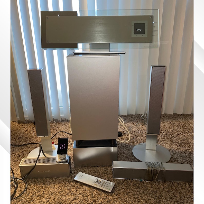 May include: A silver home theater system with a DVD player, subwoofer, two tall speakers, and a soundbar. The system includes a remote control and a small control panel. The speakers are on white stands, and the system is on a brown carpet.