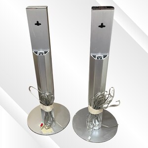 May include: Two silver-colored tower speakers with circular bases. Each speaker has a rectangular column and a bundle of gray wires secured with tape. The speakers are designed for audio systems.