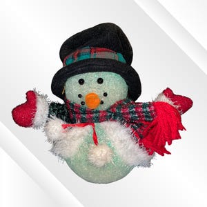 May include: A snowman figurine with a textured, light green body, wearing a black top hat with a plaid band, a matching plaid scarf, and red mittens. The snowman has a bright orange carrot-shaped nose, black bead eyes, and a white fuzzy trim.