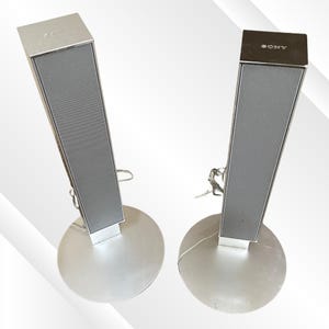 May include: Two silver Sony speakers with a rectangular design and a mesh front. The speakers are mounted on round silver bases. The word "SONY" is visible on the top of one speaker.