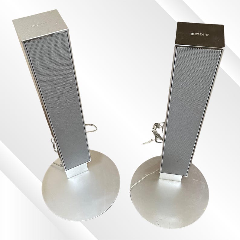 May include: Two silver Sony speakers with a rectangular design and a mesh front. The speakers are mounted on round silver bases. The word "SONY" is visible on the top of one speaker.