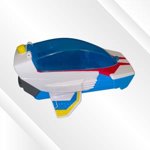 May include: A white, blue, and red plastic toy vehicle featuring a translucent blue cockpit canopy, a small yellow button on the rear, and side wings with yellow and blue accents. The design includes molded structural details and a streamlined shape.