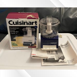 May include: A Cuisinart Mini-Prep food processor with a blue base and clear bowl, alongside its original box and instruction booklet. The box features the product name and a picture of the processor. The processor has two speed settings.