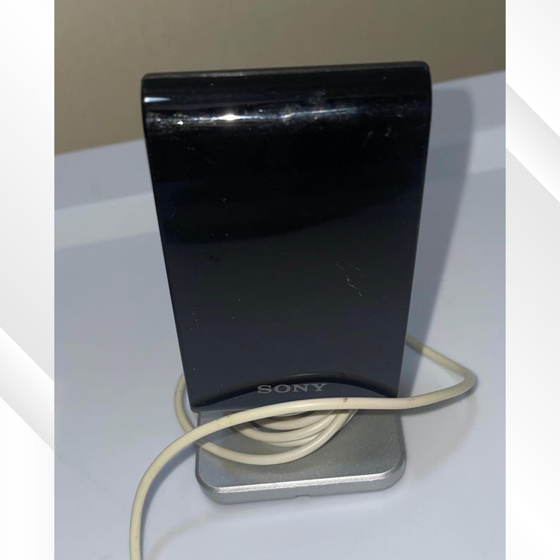 May include: A black speaker with a silver base and a white cord. The speaker has the brand name "SONY" printed on the front. The speaker is rectangular and has a glossy finish.