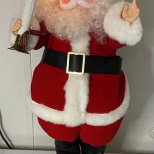 May include: Animated Santa Claus figure in a red suit with white trim, black belt, and boots. Santa holds a candle with a flickering light and has a friendly expression. The figure is on a black base.