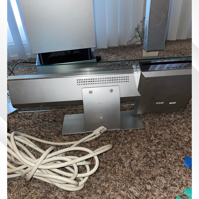 May include: Silver-colored home theater system with a glass shelf. The system includes a soundbar, two speakers, and a subwoofer. A white cable is coiled on the floor in front of the system.