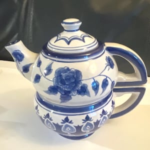 May include: A white and blue ceramic teapot with a matching cup. The teapot features a floral design with blue accents. The handle and spout are also blue. The cup is attached to the teapot. The teapot is a decorative item.