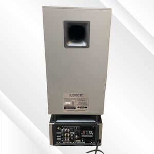May include: A silver S-master digital amplifier with a black speaker port. The back panel features multiple input and output ports, along with a power cord. The unit has a rectangular shape.