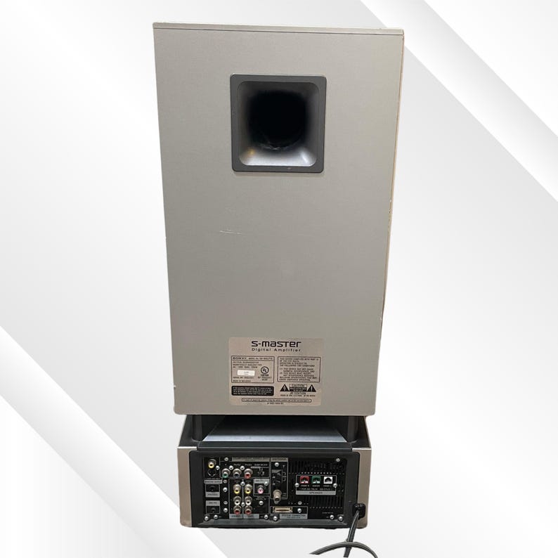 May include: A silver S-master digital amplifier with a black speaker port. The back panel features multiple input and output ports, along with a power cord. The unit has a rectangular shape.