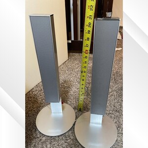May include: Two silver tower speakers with a rectangular design and a mesh top. Each speaker is mounted on a round silver base. A measuring tape is placed between the speakers, showing a measurement of approximately 18 inches.