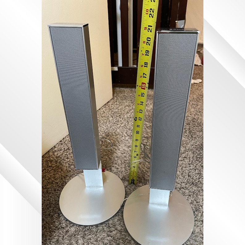 May include: Two silver tower speakers with a rectangular design and a mesh top. Each speaker is mounted on a round silver base. A measuring tape is placed between the speakers, showing a measurement of approximately 18 inches.