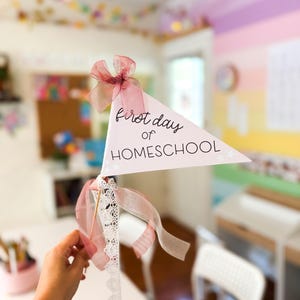 Pode incluir: Uma flâmula triangular num pau de madeira com o texto "first day of HOMESCHOOL" em letras pretas. A bandeira é decorada com um laço de organza rosa seco no topo e uma fita de renda branca atada na base do pau.