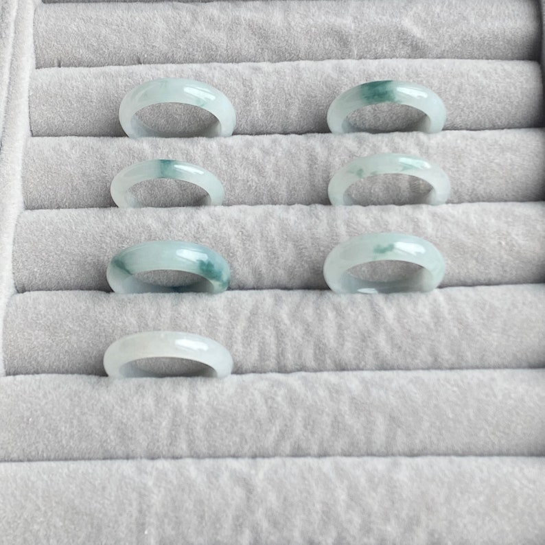 May include: Seven jade rings in a ring sizing display. The rings are a light green color with darker green veins.