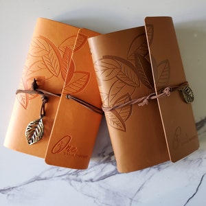 May include: Two brown leather journals with a leaf design and a gold leaf charm. The journal on the left has the text "One peace therapy" on the front.