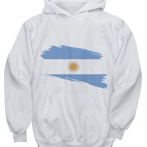 以下が含まれることがあります： 白地に青と白のアルゼンチン国旗のデザインが施されたフード付きスウェットシャツ。国旗は筆で描いたようなスタイルで描かれています。