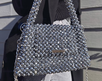 May include: A silver beaded handbag with a top handle and a chain strap. The bag is made of small, round beads that are tightly woven together.