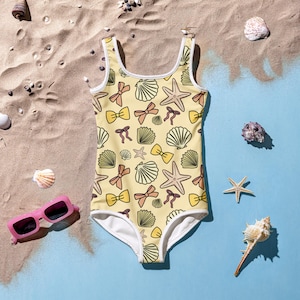 May include: A yellow one-piece swimsuit with a pattern of seashells, starfish, and bows. The swimsuit has white trim and is set on a beach scene with pink sunglasses and seashells.