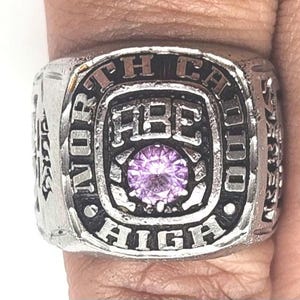 May include: Silver-toned ring with the words "NORTH CAL HIGH" and "ABE" engraved on it. The ring features a central, round, light purple gemstone. The ring is worn on a finger.