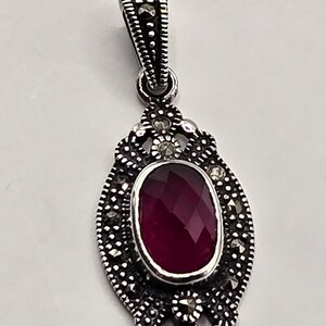 May include: A silver pendant with an oval, faceted, deep red gemstone. The pendant has an ornate, filigree design with small, round accents. The bail is also detailed with a similar pattern. The jewelry is likely a necklace pendant.