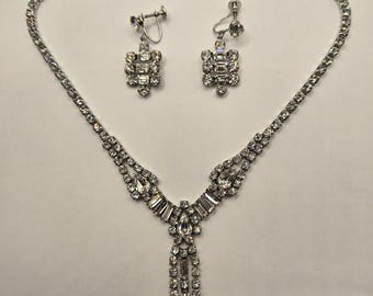 Op de afbeelding: Een zilverkleurige sieradenset met een ketting en bijpassende oorbellen. De ketting heeft een fijne ketting met een centrale hanger van heldere kristallen. De oorbellen zijn vierkant en hebben vergelijkbare kristallen details. Geschikt voor formele gelegenheden.