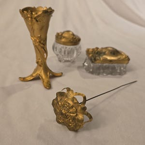 May include: An assortment of antique gold-toned vanity items. Includes a small vase, two glass jars with gold lids, and a decorative pin. The items have ornate floral and scrollwork designs, suggesting a vintage aesthetic.