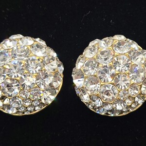 Puede incluir: Un par de pendientes de botón redondos y brillantes. Cada pendiente está cubierto de numerosos cristales transparentes y facetados, engastados en una base de metal dorado. Los cristales captan la luz, creando un efecto deslumbrante. Fondo oscuro.