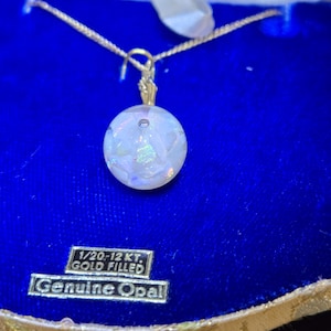 Puede incluir: Un collar de oro relleno con un colgante redondo de ópalo. El colgante es blanco con destellos iridiscentes. El collar está sobre un fondo de terciopelo azul. La etiqueta dice "1/20-12 Kt. Gold Filled Genuine Opal."