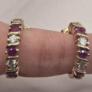May include: A pair of gold hoop earrings. Each earring features alternating round, deep red gemstones and clear, sparkling stones. The gemstones are set in a twisted gold design.