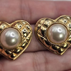 May include: A pair of gold-coloured heart-shaped earrings with a quilted pattern. Each earring has a large, round, off-white pearl in the centre. The earrings are displayed against a light background.