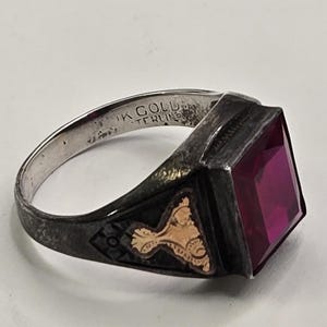 Puede incluir: Un anillo vintage con una gema rectangular de color rojo intenso. El anillo tiene una banda plateada con el texto "10K GOLD STERLING" y un engaste negro con detalles de filigrana dorada. La gema es el punto focal del anillo.
