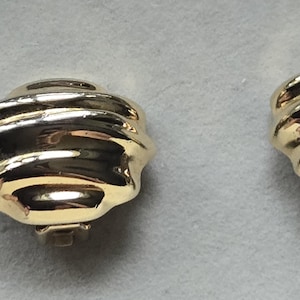May include: A pair of gold-coloured clip-on earrings. Each earring features a ridged, rounded design with a shiny, reflective surface. The earrings are set against a neutral background.