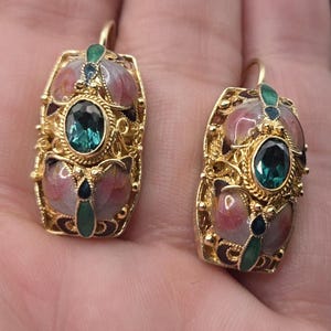 May include: A pair of ornate gold-toned earrings. Each earring features a central oval teal-colored stone, surrounded by intricate gold filigree and pink and green enamel details. The earrings have a hook closure.