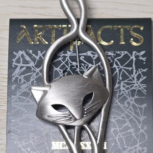 Peut inclure: Une broche en métal argenté représentant un chat stylisé avec une queue arquée. La broche est fixée sur une carte de présentation noire avec le texte doré ARTIFACTS et MC XXI, ornée d'un motif de lignes abstraites de couleur blanche.