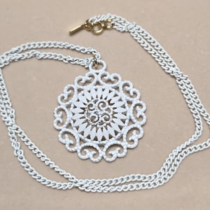 Op de afbeelding: Een witte hangerketting met een gedetailleerd, opengewerkt ontwerp. De hanger heeft een cirkelvorm met een bloemmotief. De ketting is een dubbele streng van wit metaal. De sluiting is goudkleurig.