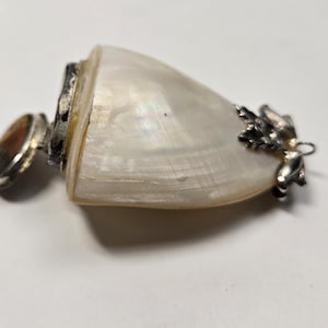 May include: A pearlescent white shell pendant with silver-colored metal accents. The pendant has a rounded shape with a silver-colored bail and a small, round, brown stone. The shell has a natural, iridescent sheen.