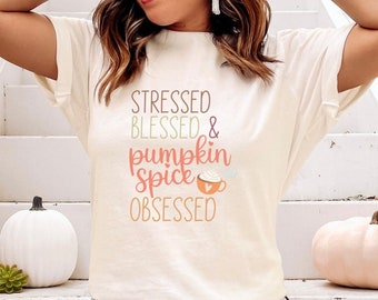 May include: A woman wearing a white t-shirt with the text "Stressed Blessed & Pumpkin Spice Obsessed" in colorful lettering. She is sitting on a set of white stairs with pumpkins around her.