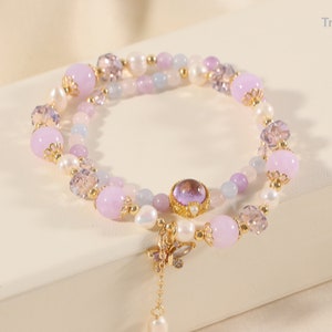 May include: A delicate bracelet featuring a combination of lavender, white, and clear beads. The bracelet includes a butterfly charm and a dangling pearl. The design incorporates gold accents and a central purple gemstone.