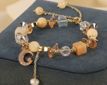 May include: A gold bracelet with a moon and star charm, featuring a variety of beige and clear beads. The bracelet is displayed on a blue velvet box.