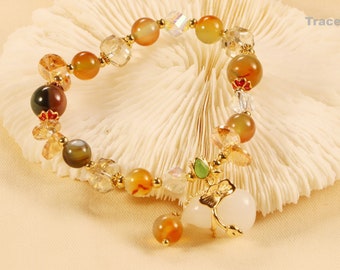 Pode incluir: Um bracelete dourado com um amuleto de pêssego. O bracelete é feito com contas de várias cores, incluindo transparente, marrom, verde e laranja.