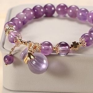 May include: A purple amethyst gemstone bracelet with a gold charm in the shape of a pouch. The bracelet has a delicate gold chain with a small amethyst bead at the end.
