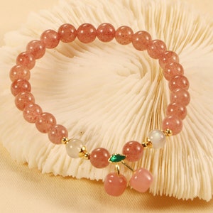 Op de afbeelding: Een roze en witte kralen armband met een gouden bedel van twee roze kersen met een groen blaadje. De armband ligt op een wit koraalachtig oppervlak.
