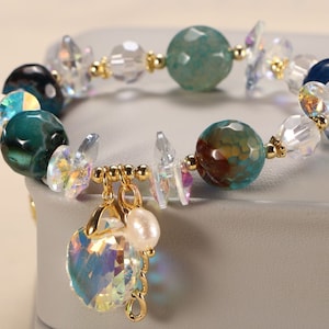 Puede incluir: Un brazalete de cuentas de ágata turquesa, azul y marrón con detalles dorados y un dije de cristal. El brazalete tiene una perla y un colgante de cristal.