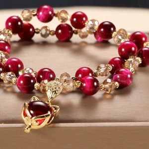 Könnte beinhalten: Ein doppelreihiges Armband mit roten Tigeraugenperlen und goldenen Akzenten. Das Armband hat einen goldenen Fuchs-Anhänger mit einem roten Edelstein.