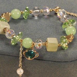 Könnte beinhalten: Ein grünes und goldenes Perlenarmband mit einem Anhänger in Form eines Planeten mit einem grünen Edelstein. Das Armband besteht aus verschiedenen Perlen, darunter grüne Glasperlen, klare Glasperlen und goldene Metallperlen.