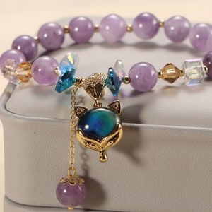 May include: A purple amethyst beaded bracelet with a gold fox charm and a dangling amethyst bead. The bracelet is on a white background.