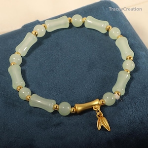 May include: A light green jade bracelet with gold accents and a gold charm in the shape of two leaves. The bracelet is made of round beads and rectangular beads.