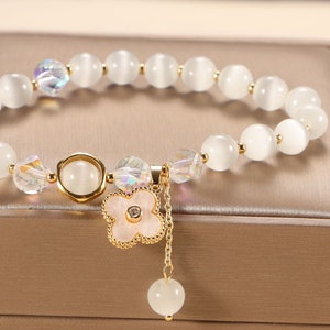 May include: White gemstone bracelet with a gold flower charm and a small white gemstone dangling from a chain. The bracelet is displayed in a beige box.