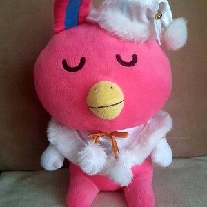 Puede incluir: Un juguete de peluche rosa con ribete de piel blanca y un pico amarillo. El juguete lleva una gorra de dormir blanca con un pompón blanco y una cinta dorada alrededor del cuello.