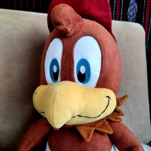 May include: A brown plush toy bird wearing a red baseball cap. The bird has a yellow beak, blue eyes, and a white belly.